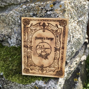 Puede incluir: Una placa rectangular de madera con acabado en tono beige claro que presenta bordes y texto decorativos grabados con l&aacute;ser en color marr&oacute;n oscuro. El centro muestra el texto Jhotun's Forge by The Dungeon of Rose, rodeado de un marco.