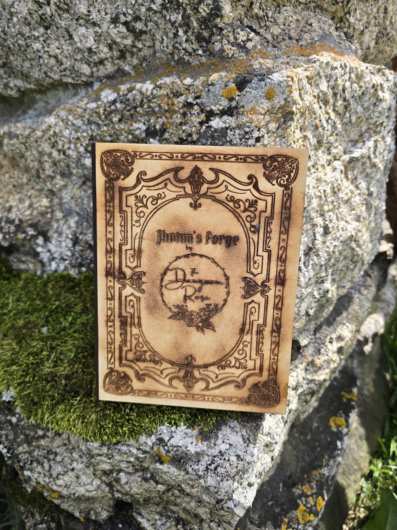 Puede incluir: Una placa rectangular de madera con acabado en tono beige claro que presenta bordes y texto decorativos grabados con l&aacute;ser en color marr&oacute;n oscuro. El centro muestra el texto Jhotun's Forge by The Dungeon of Rose, rodeado de un marco.