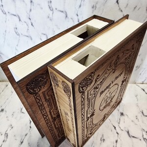 Puede incluir: Dos cajas rectangulares de madera dise&ntilde;adas para parecer libros con interiores huecos. La cubierta frontal de la caja m&aacute;s clara presenta bordes decorativos grabados con l&aacute;ser y el texto "Jotun's Forge" con un emblema circular central.