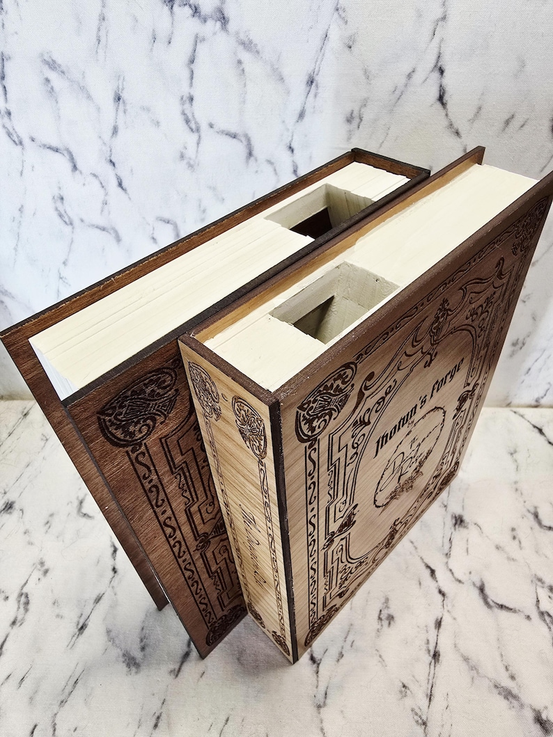 Puede incluir: Dos cajas rectangulares de madera dise&ntilde;adas para parecer libros con interiores huecos. La cubierta frontal de la caja m&aacute;s clara presenta bordes decorativos grabados con l&aacute;ser y el texto "Jotun's Forge" con un emblema circular central.