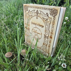 Puede incluir: Una cubierta de libro de madera clara con bordes decorativos grabados con l&aacute;ser y el texto Jotun's Forge by The Dungeon Rose. La cubierta rectangular presenta un emblema circular central y dise&ntilde;os intrincados de volutas en las esquinas.