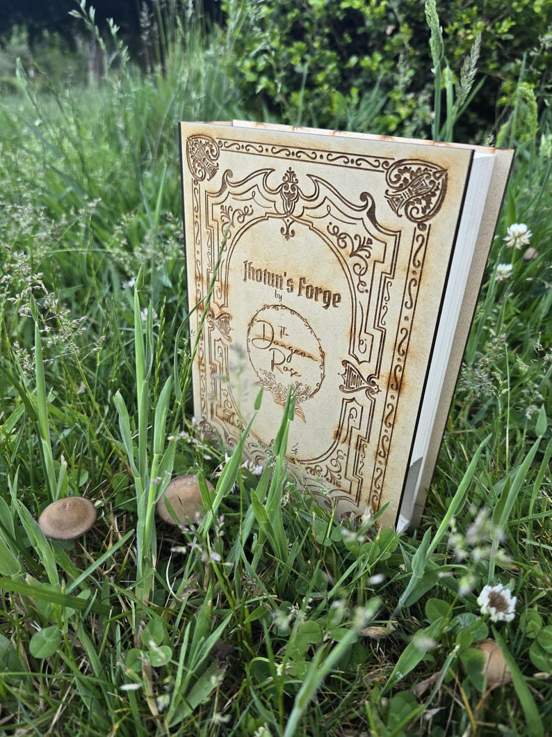 Puede incluir: Una cubierta de libro de madera clara con bordes decorativos grabados con l&aacute;ser y el texto Jotun's Forge by The Dungeon Rose. La cubierta rectangular presenta un emblema circular central y dise&ntilde;os intrincados de volutas en las esquinas.