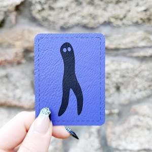 May include: A purple card with a black silhouette of a creature with two eyes. The creature has long legs and arms.