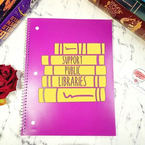 May include: A purple spiral notebook with a yellow graphic of stacked books that says "Support Public Libraries".