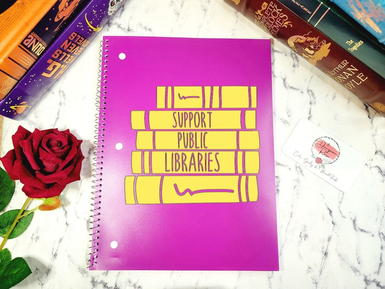 May include: A purple spiral notebook with a yellow graphic of stacked books that says "Support Public Libraries".