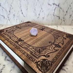 Puede incluir: Una caja rectangular de madera con acabado marr&oacute;n oscuro y patrones de filigrana grabados con l&aacute;ser en la tapa. En el centro de la tapa descansa un dado de veinte caras de color violeta claro transl&uacute;cido con n&uacute;meros violeta oscuro.
