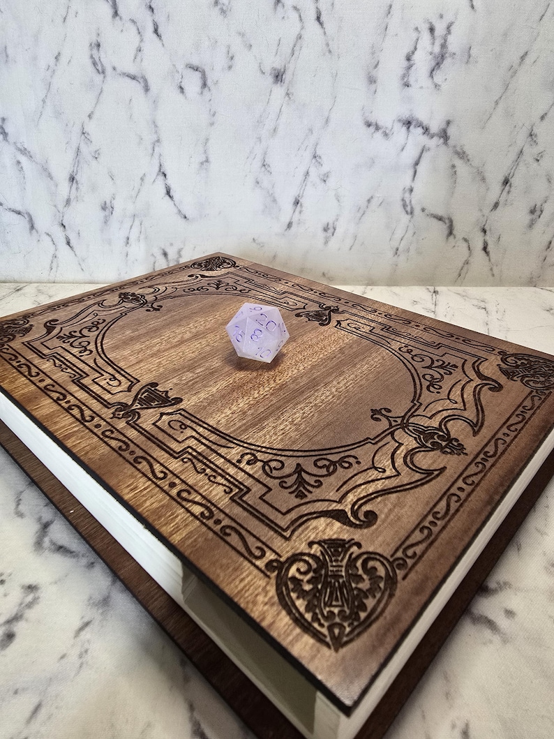 Puede incluir: Una caja rectangular de madera con acabado marr&oacute;n oscuro y patrones de filigrana grabados con l&aacute;ser en la tapa. En el centro de la tapa descansa un dado de veinte caras de color violeta claro transl&uacute;cido con n&uacute;meros violeta oscuro.