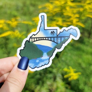 May include: A sticker of the state of West Virginia with a bridge and a flying bird silhouette. The sticker is white with a blue sky, green mountains, and a brown bridge.