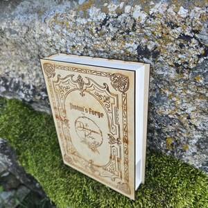 Puede incluir: Una caja rectangular de madera dise&ntilde;ada como un libro con acabado en tono bronceado claro y bordes decorativos grabados a l&aacute;ser en marr&oacute;n oscuro. La portada presenta el texto Jhotun's Forge by The Dragon Rose con un emblema floral circular.
