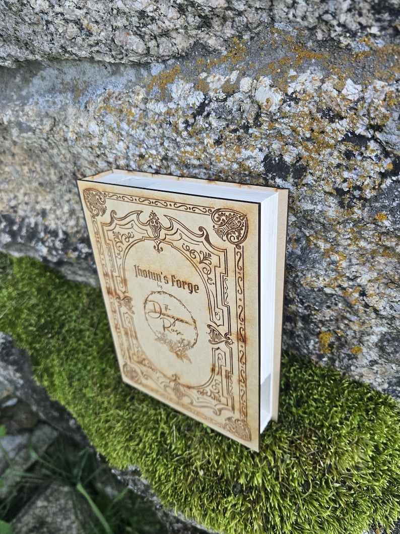 Puede incluir: Una caja rectangular de madera dise&ntilde;ada como un libro con acabado en tono bronceado claro y bordes decorativos grabados a l&aacute;ser en marr&oacute;n oscuro. La portada presenta el texto Jhotun's Forge by The Dragon Rose con un emblema floral circular.
