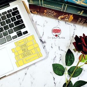 May include: A yellow sticker with the text "Support Public Libraries" on a silver laptop. The sticker is shaped like a stack of books. A red rose and a business card are on a white marble surface.