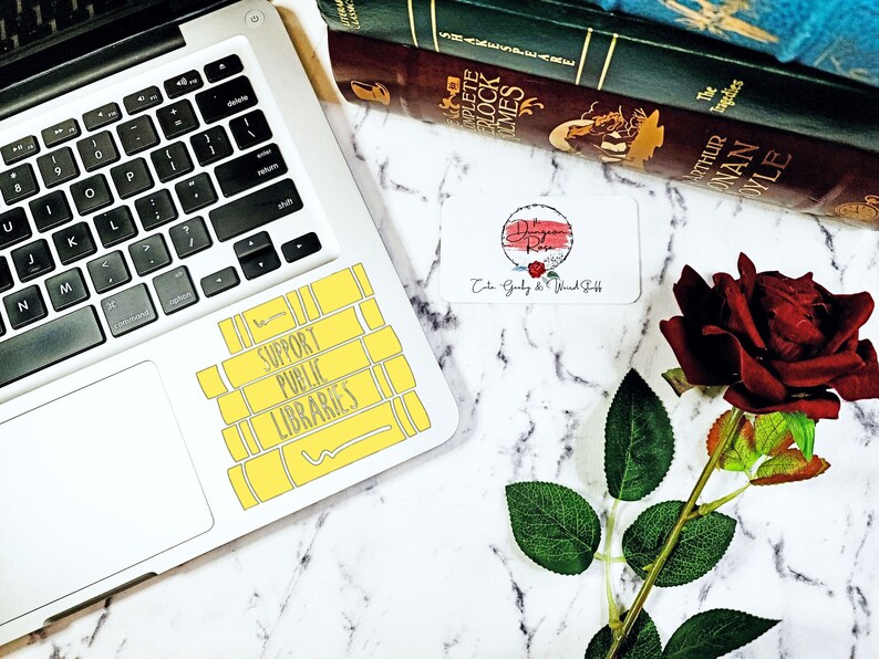 May include: A yellow sticker with the text "Support Public Libraries" on a silver laptop. The sticker is shaped like a stack of books. A red rose and a business card are on a white marble surface.