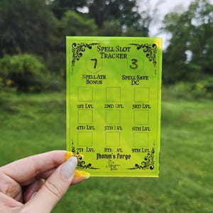 May include: A rectangular, translucent neon yellow acrylic spell slot tracker card featuring black engraved text and grid boxes. The card displays headers for spell attack bonus, spell save DC, and levels one through nine, with the text Jhotun's Forge.