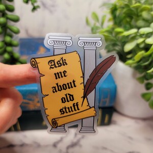 May include: A clear sticker with a yellow scroll that says "Ask me about old stuff". The scroll is between two white columns with a brown feather quill.