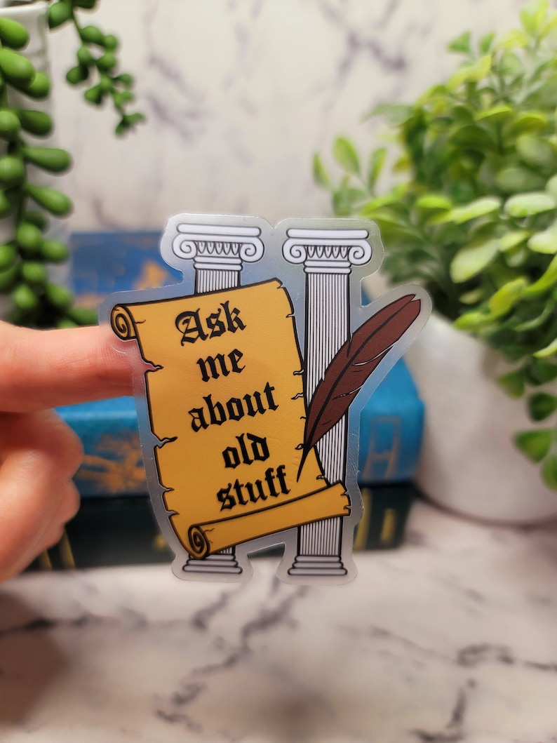 May include: A clear sticker with a yellow scroll that says "Ask me about old stuff". The scroll is between two white columns with a brown feather quill.