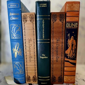 Puede incluir: Una colecci&oacute;n de cinco libros colocados verticalmente. De izquierda a derecha: un Robinson Crusoe azul, un The Dungeon Rose de madera clara, un Shakespeare The Tragedies verde oscuro, un The Dungeon Rose de madera oscura y un Dune naranja de Frank Herbert.