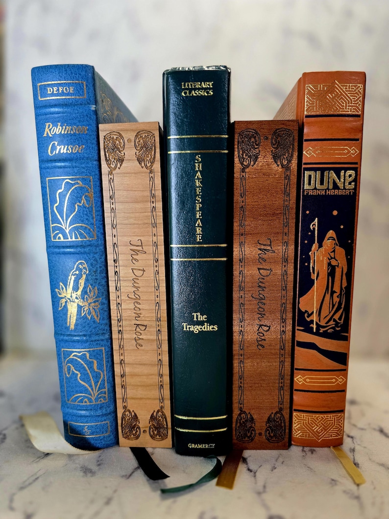 Puede incluir: Una colecci&oacute;n de cinco libros colocados verticalmente. De izquierda a derecha: un Robinson Crusoe azul, un The Dungeon Rose de madera clara, un Shakespeare The Tragedies verde oscuro, un The Dungeon Rose de madera oscura y un Dune naranja de Frank Herbert.