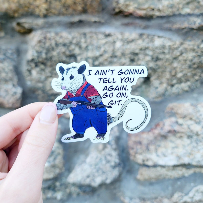 May include: A white opossum wearing overalls and holding a shotgun. The opossum is looking to the left. The text "I ain't gonna tell you again. Go on, git." is written above the opossum.