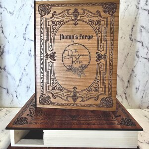 Puede incluir: Una caja de madera con forma de libro con una cubierta de color marr&oacute;n claro que presenta bordes ornamentados grabados con l&aacute;ser y el texto Jhotun's Forge by The Dungeon Rose. La base es de madera oscura con p&aacute;ginas blancas visibles al costado.