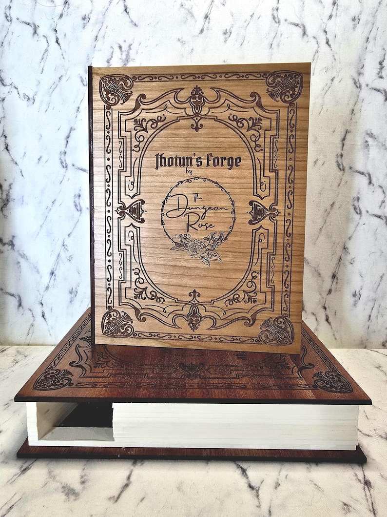 Puede incluir: Una caja de madera con forma de libro con una cubierta de color marr&oacute;n claro que presenta bordes ornamentados grabados con l&aacute;ser y el texto Jhotun's Forge by The Dungeon Rose. La base es de madera oscura con p&aacute;ginas blancas visibles al costado.
