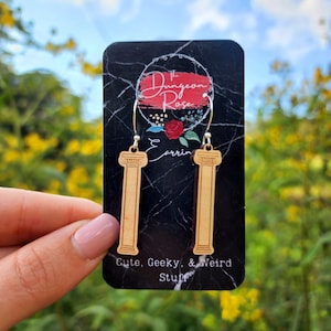 May include: A pair of wooden earrings shaped like columns with a silver hoop. The earrings are displayed on a black card with the text "The Dungeon Rose Earrings Cute, Geeky, & Weird Stuff".
