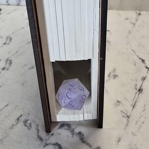 Puede incluir: Un libro ahuecado con un exterior marr&oacute;n oscuro y p&aacute;ginas blancas, que presenta una secci&oacute;n recortada en el centro. Dentro del recorte se encuentra un dado de veinte caras de color violeta claro con n&uacute;meros violeta oscuro, incluyendo un 20, 14, 10, 8 y 5 visibles.