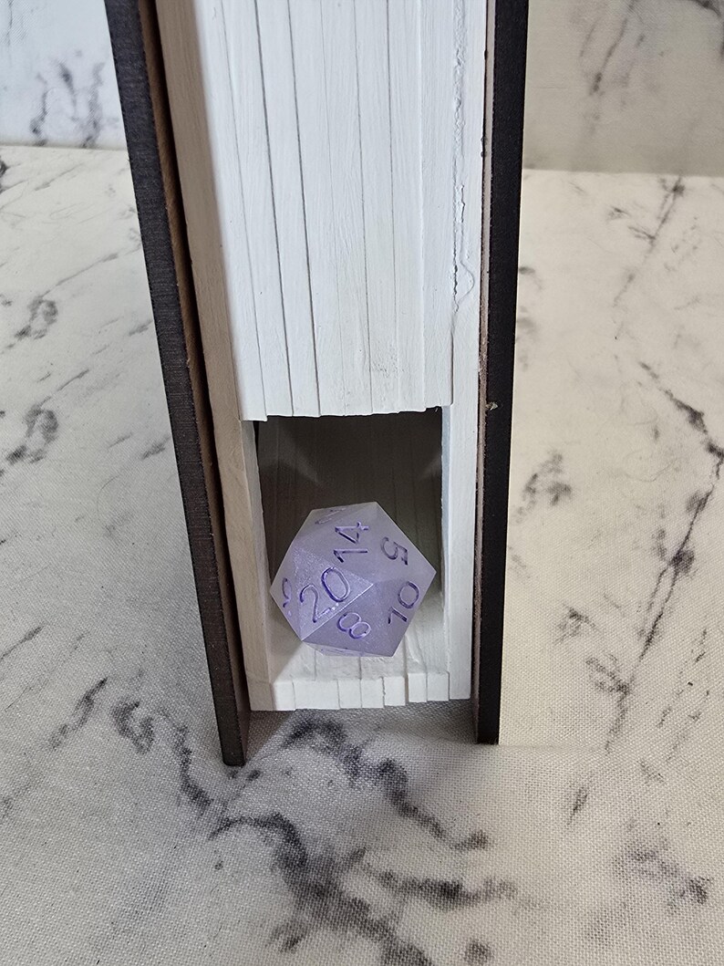 Puede incluir: Un libro ahuecado con un exterior marr&oacute;n oscuro y p&aacute;ginas blancas, que presenta una secci&oacute;n recortada en el centro. Dentro del recorte se encuentra un dado de veinte caras de color violeta claro con n&uacute;meros violeta oscuro, incluyendo un 20, 14, 10, 8 y 5 visibles.