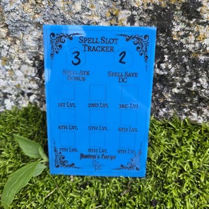May include: A rectangular blue acrylic spell slot tracker featuring black engraved text and decorative flourishes. The board includes sections for Spell Slot Tracker, Spell Atk Bonus, Spell Save DC, and levels 1st through 9th, with Jhotun's Forge branding.