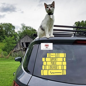 May include: A white and gray cat sits on the back of a gray car. A yellow sticker on the car reads "Support Public Libraries" with a stack of books illustrated in yellow.