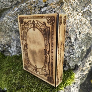 Puede incluir: Una caja rectangular de madera con acabado en tono bronceado claro y patrones de borde ornamentados grabados con l&aacute;ser en marr&oacute;n oscuro en la tapa. El panel lateral presenta el texto The Dungeon Rosa grabado en letra cursiva oscura.