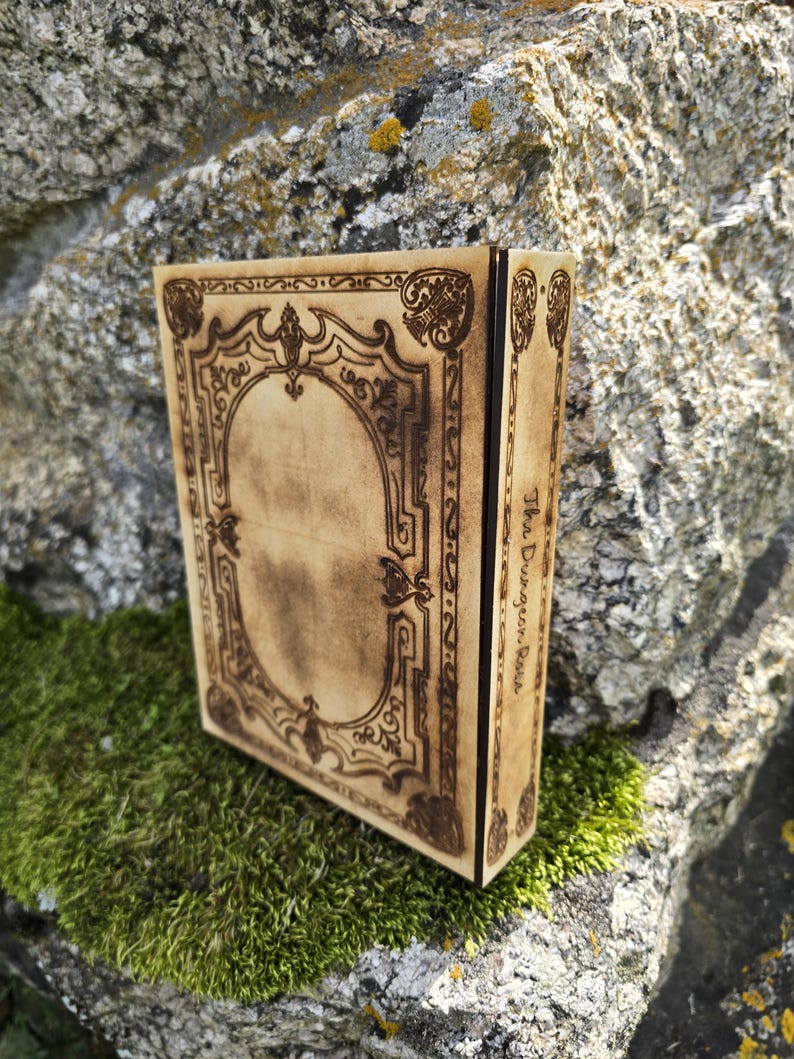 Puede incluir: Una caja rectangular de madera con acabado en tono bronceado claro y patrones de borde ornamentados grabados con l&aacute;ser en marr&oacute;n oscuro en la tapa. El panel lateral presenta el texto The Dungeon Rosa grabado en letra cursiva oscura.