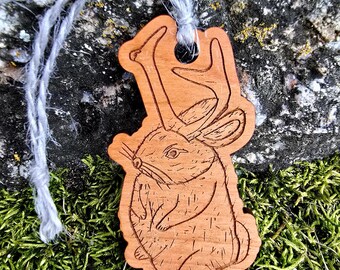 May include: A wooden ornament of a rabbit with antlers. The rabbit is brown and has a detailed design. The ornament is hanging from a white string.