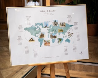 May include: A framed world map seating chart on a wooden easel. The map is light blue and features watercolor illustrations of landmarks like the Eiffel Tower and the Colosseum. Names are listed under city names. The frame is gold.