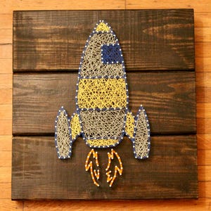 May include: A string art rocket ship with yellow, blue, and gray string on a dark brown wooden background.