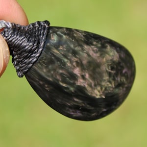 May include: Close-up of a teardrop-shaped pendant. The pendant is a dark, swirling mix of black, gray, and hints of pink. It is attached to a gray cord with a woven black cap. The background is a soft green.