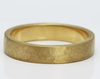 May include: A gold band ring featuring a textured, hammered finish on the exterior surface. The ring has a flat profile and a consistent width throughout, showcasing a matte, brushed appearance with subtle indentations across the metal.