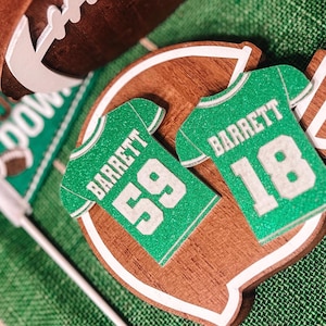 May include: A collection of football-themed decorations, including a football, a touchdown flag, and wooden cutouts shaped like jerseys and a field. The jerseys are green with the name "Barrett" and the numbers 59 and 18.