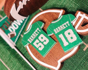 May include: A collection of football-themed decorations, including a football, a touchdown flag, and wooden cutouts shaped like jerseys and a field. The jerseys are green with the name "Barrett" and the numbers 59 and 18.