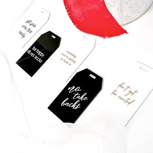 Op de afbeelding: Vier bagagelabeltjes met gouden letters op een zwarte en witte achtergrond. De tekst op de labeltjes luidt: "get your wow face ready", "be happy it's not socks", "warning you're going to hate it", "no take backs", en "don't get too excited".