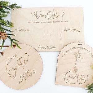 Op de afbeelding: Drie houten borden voor Kerstmis. Het eerste bord is rond en heeft de tekst "Lieve Kerstman, stop hier alstublieft!" Het tweede bord is rechthoekig en heeft de tekst "Lieve Kerstman, bedankt voor het bezoeken van ons huis vanavond! Kerstman's melk Koekjes voor de Kerstman Voor de rendieren". Het derde bord is een boog met een sleutelgat en de tekst "Voor ons huis zonder schoorsteen De magische sleutel van de Kerstman!"