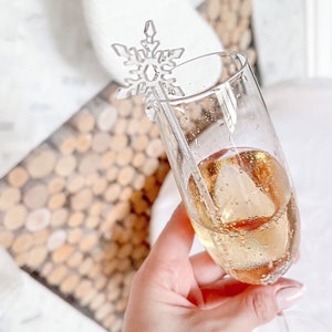 Op de afbeelding: Een heldere glazen champagnefluit met een sneeuwvlokvormige drankroerder. De fluit is gevuld met een lichtgekleurde, mousserende drank.