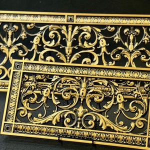 May include: Two rectangular metal vent covers featuring a raised gold-toned scrollwork pattern against a recessed black background. Each piece has a decorative border with repeating vertical lines and circular screw holes at the four corners.