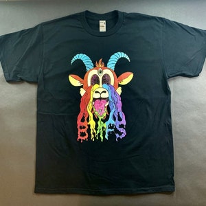 Può includere: Una t-shirt nera a maniche corte con la grafica di una testa di capra con un terzo occhio, corna blu e lingua di fuori. Un liquido color arcobaleno cola dagli occhi e dalla bocca, formando le lettere BMRS nella parte inferiore del disegno.