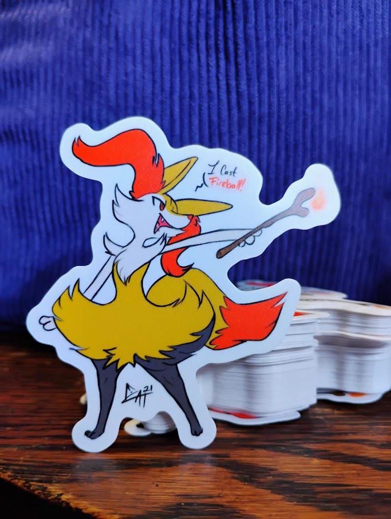 May include: A die-cut sticker featuring a cartoon fox-like character in yellow, orange, and white, holding a wand. The sticker has the text "I cast Fireball!" and the artist's signature.