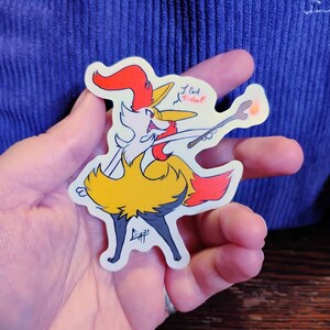 May include: A sticker featuring a cartoon fox-like character with a red and yellow color scheme, holding a wand. The text "I cast Fireball!" is written on the sticker. The sticker is held in a hand.