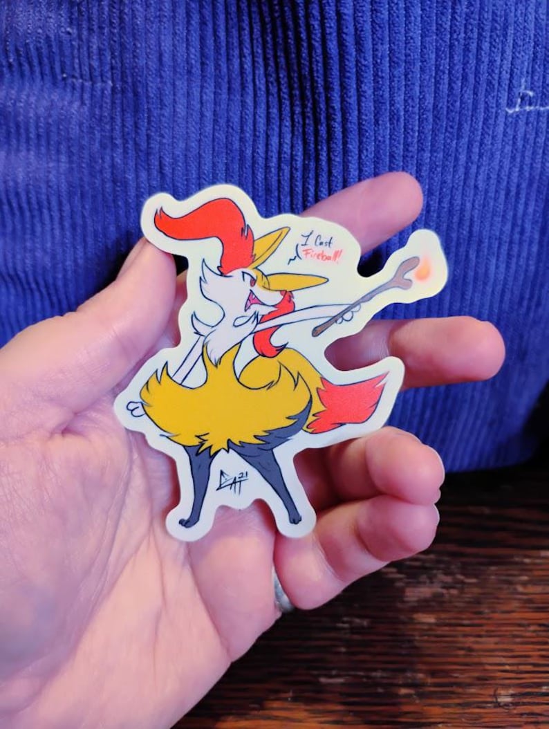 May include: A sticker featuring a cartoon fox-like character with a red and yellow color scheme, holding a wand. The text "I cast Fireball!" is written on the sticker. The sticker is held in a hand.