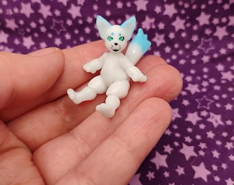 May include: A small, white figurine of a fox-like creature with blue accents on its ears and tail. The figurine has green eyes and is lying down. The background is a purple fabric with white star patterns.