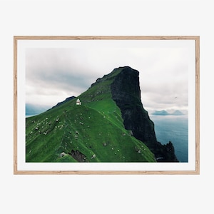 May include: A small white building with a red roof sits on a green grassy hilltop overlooking a vast ocean. The hill is part of a large, dark grey cliff face. The sky is cloudy and grey.