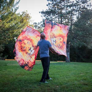 May include: A person in a grey shirt and black pants is holding two flags with lion images. The flags are red, orange, and yellow with a lion's face in the center. The person is standing in a grassy field with trees in the background.