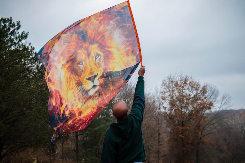 May include: A person holds a large flag with a lion's face printed on it. The lion is surrounded by flames and the flag is orange, red, and yellow.